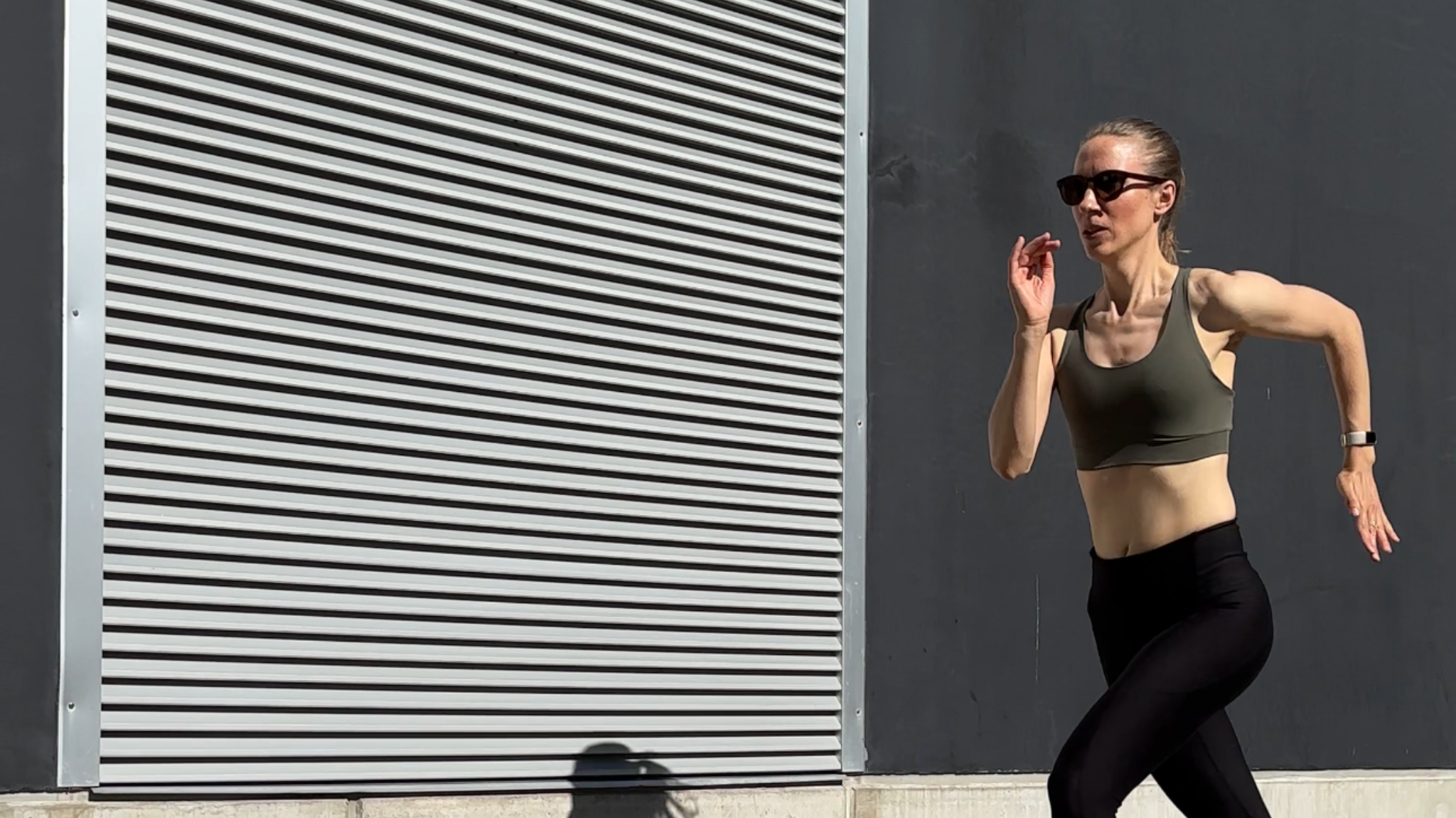 Woman running past a grey industrial wall in an olive sports bra and black leggings.