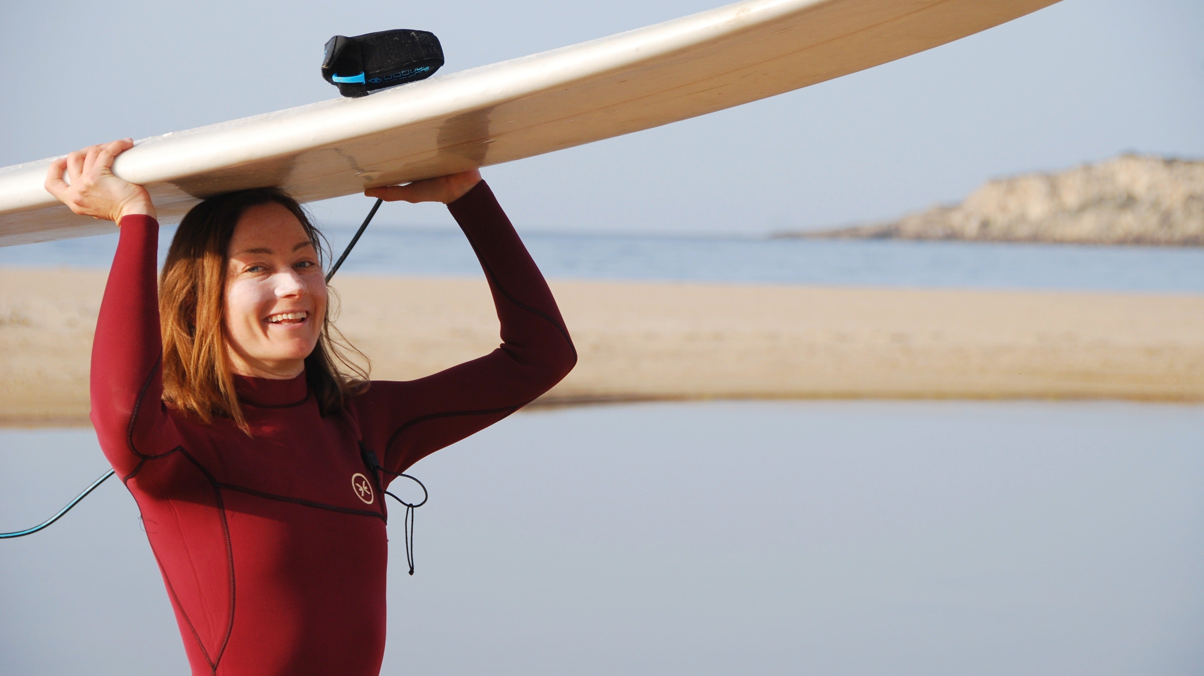 Julia Beyer from Suntribe carrying a surfboard overhead in a red wetsuit on the beach.