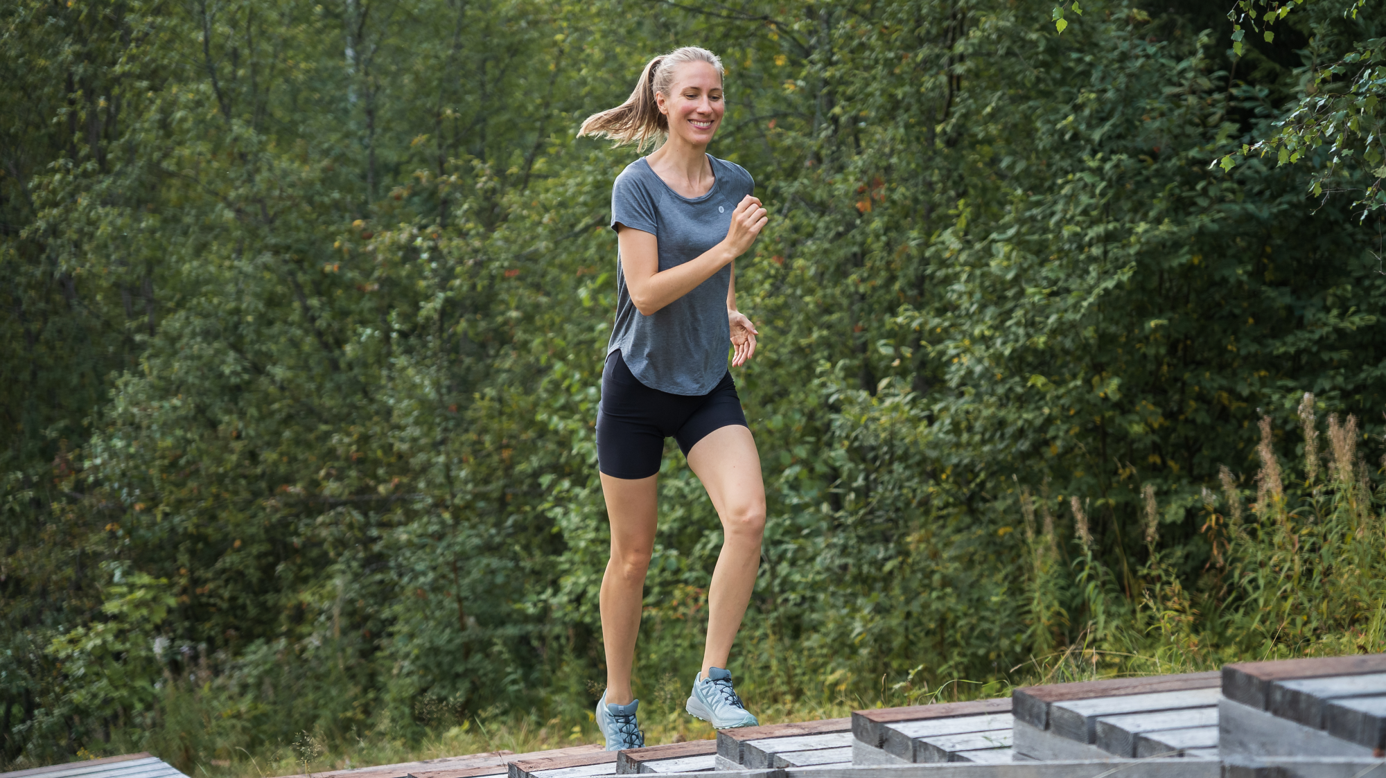Woman jogging up wooden outdoor steps in a blue Tripulse T-shirt and shorts.