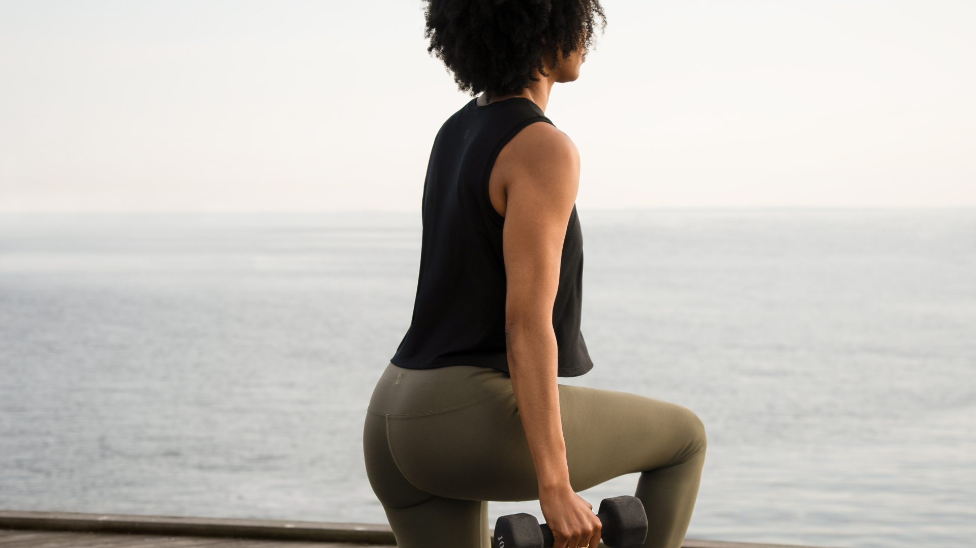Woman lunging with a dumbbell by the sea in a black top and olive leggings.