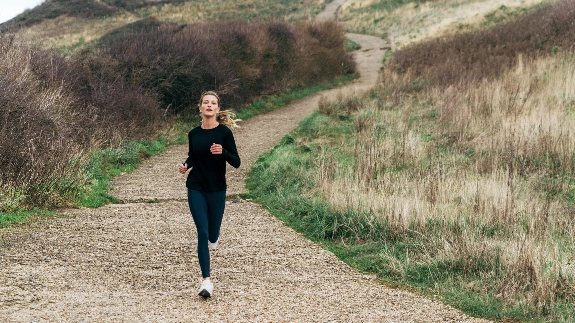 Woman running down a gravel coastal path in a black long-sleeve top and leggings.