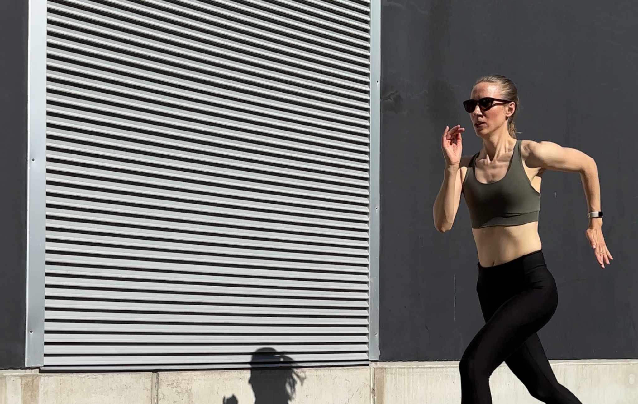 Woman running past a grey industrial wall in an olive sports bra and black leggings.