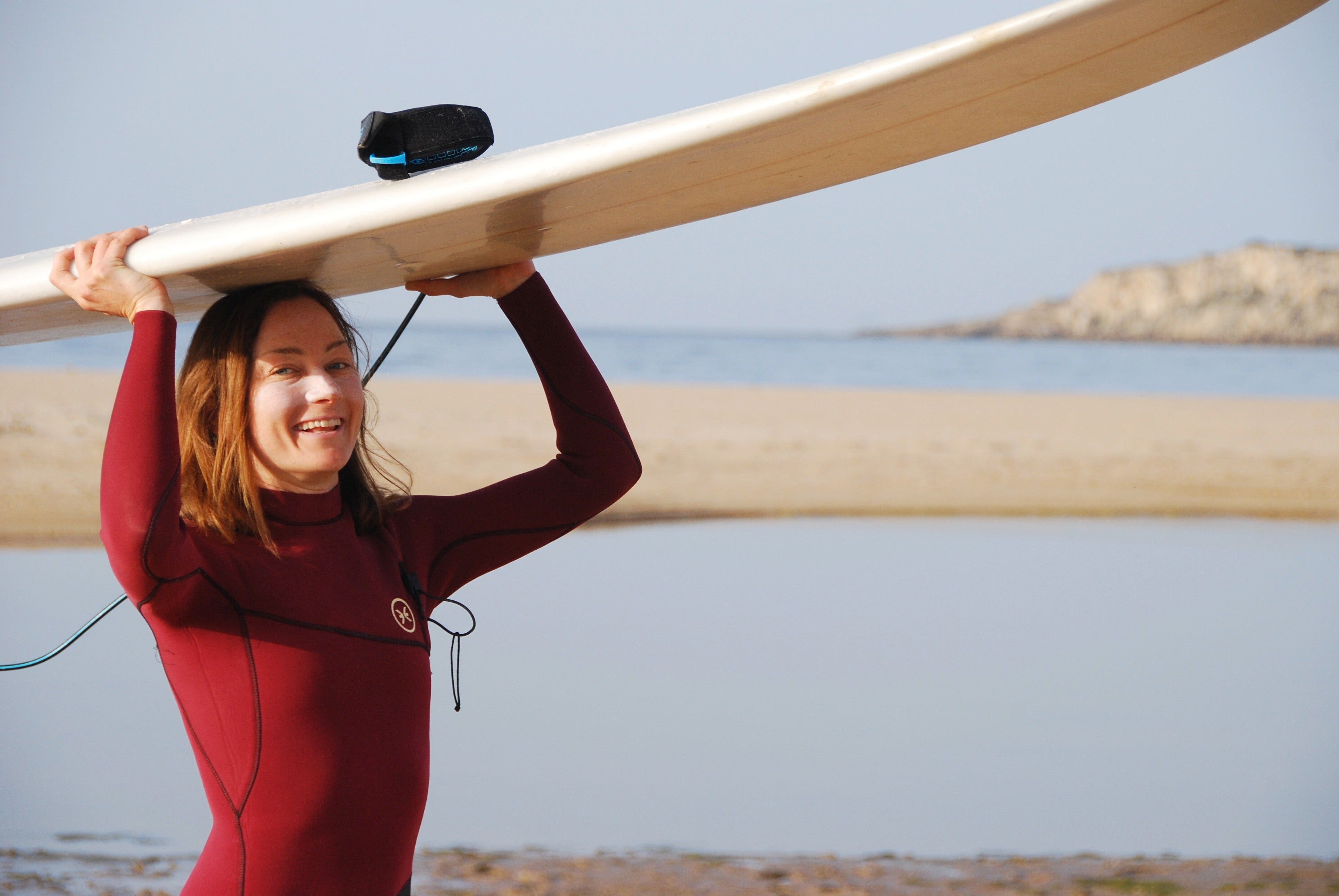 Julia Beyer from Suntribe carrying a surfboard overhead in a red wetsuit on the beach.
