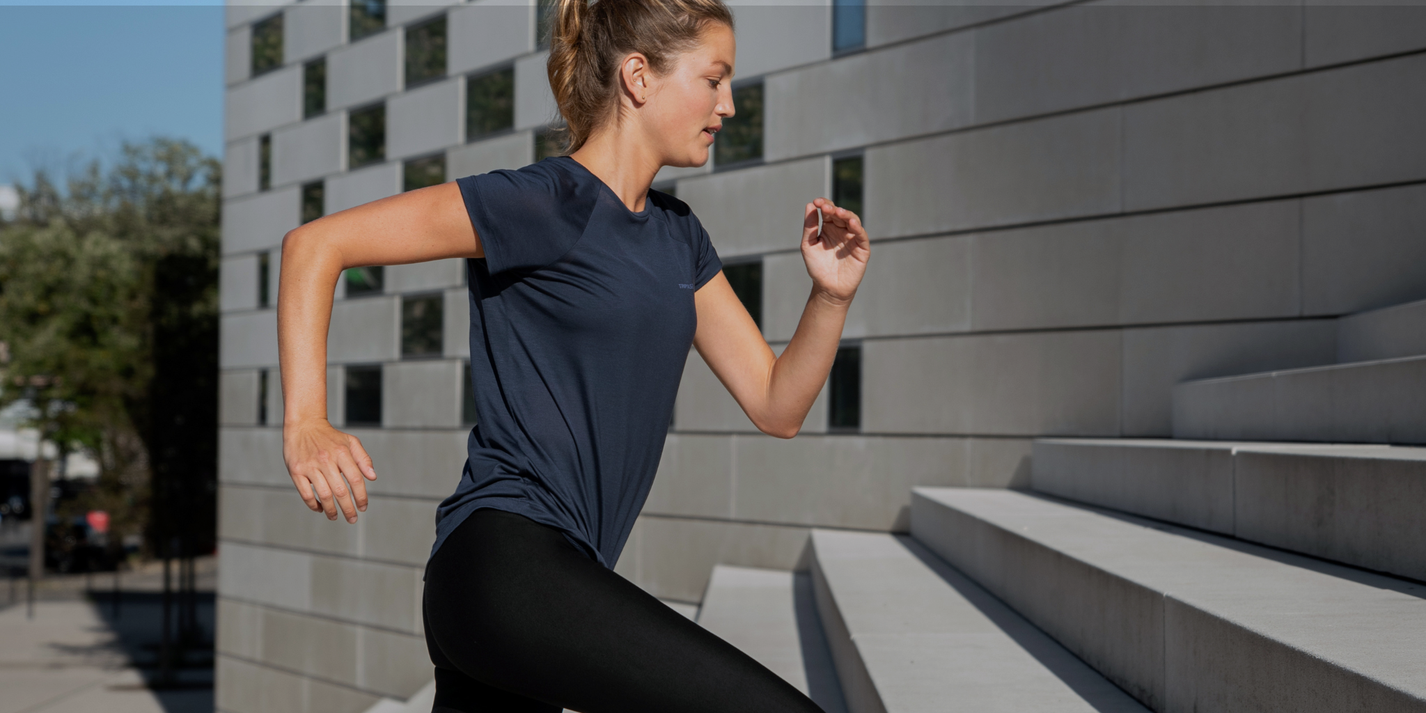 Woman running up concrete steps in a navy Tripulse T-shirt and leggings.