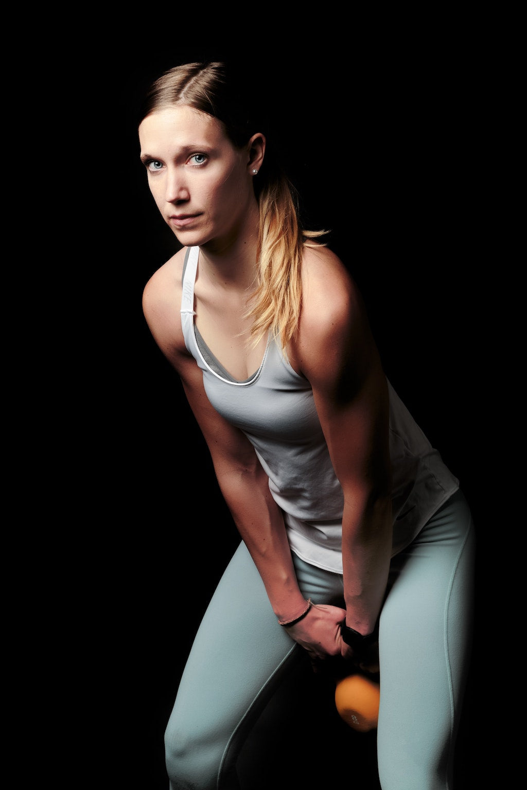 Woman in a tank top and light green leggings lifting a kettlebell against a dark background.