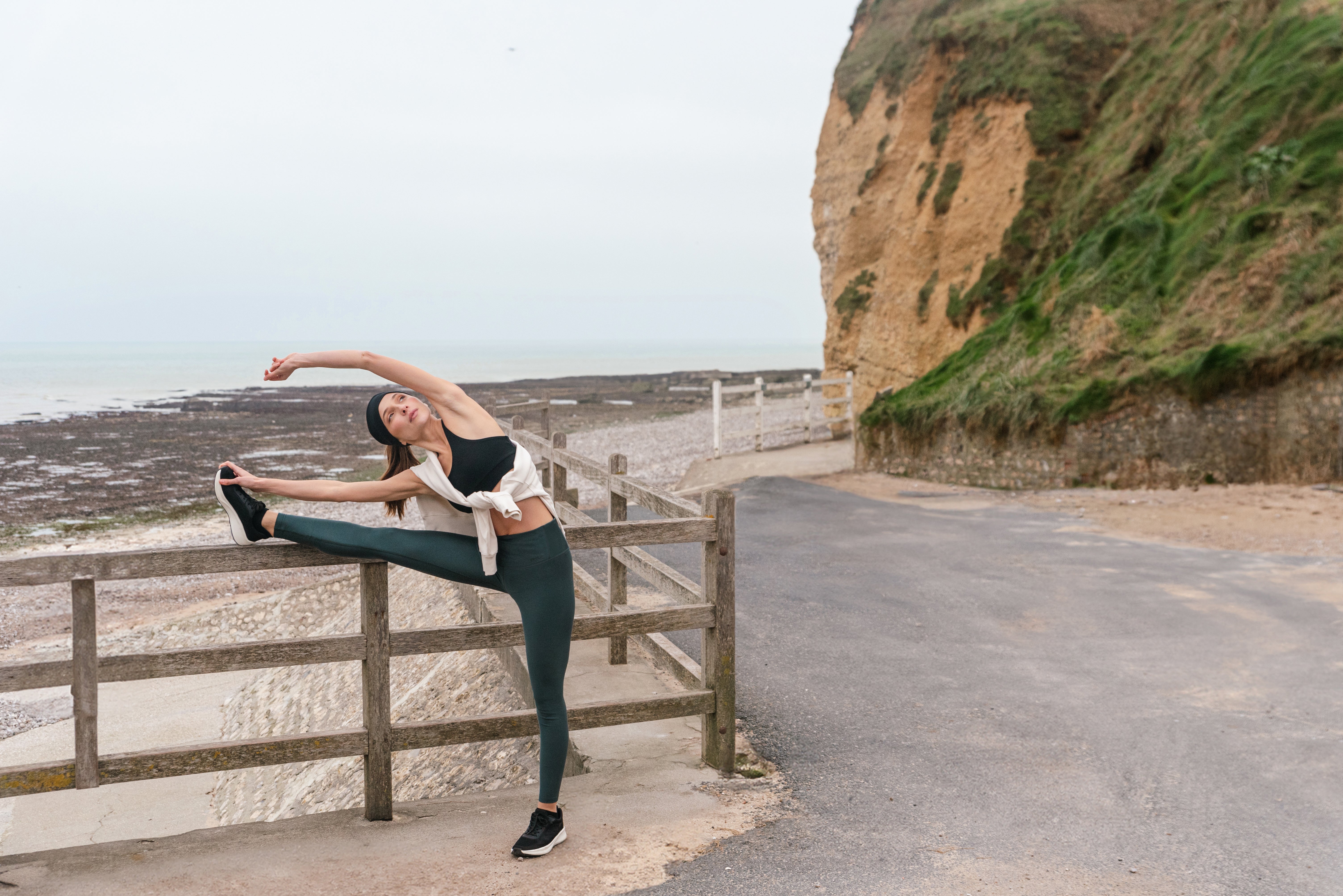 Woman stretching her leg on a fence by a coastal path in dark green Tripulse leggings.