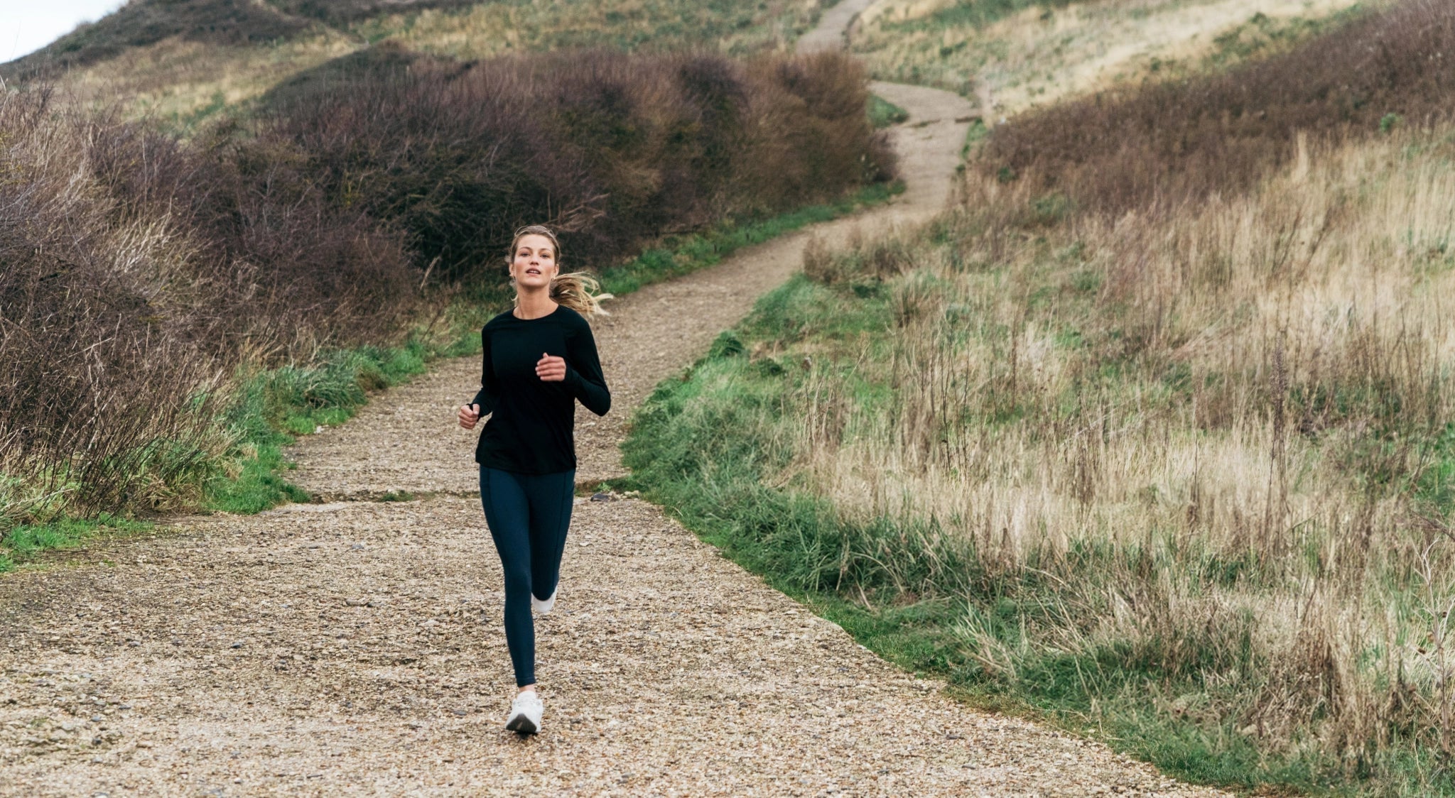 Woman running down a gravel coastal path in a black long-sleeve top and leggings.