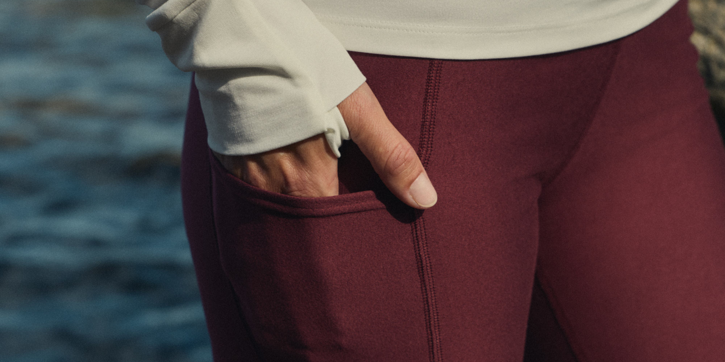 Hand in the side pocket of burgundy leggings with a cream long sleeve top, by the water